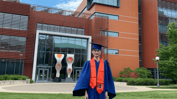 YiFei Zhu stands dressed in his graduation gown in front of the ECE building
